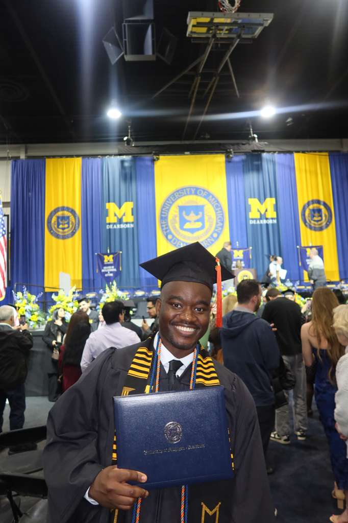 Jidechukwu Owo smiling in regalia, holding his University of Michigan-Dearborn diploma